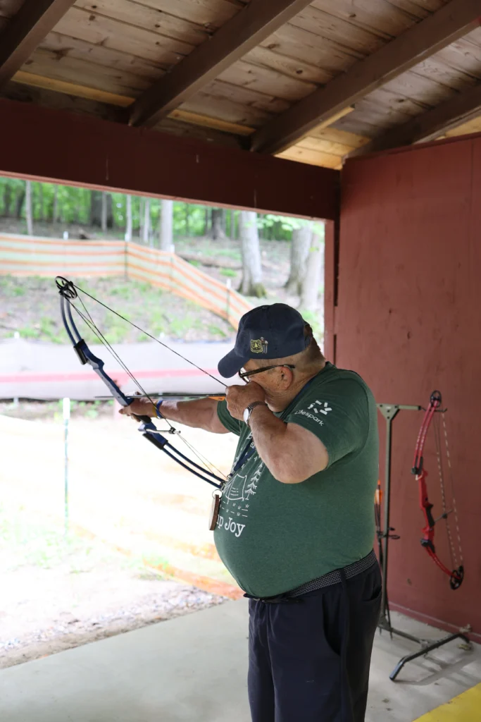 Holding a bow and aiming, a male resident of Lifespark's senior living communities participates in archery at Camp Joy.